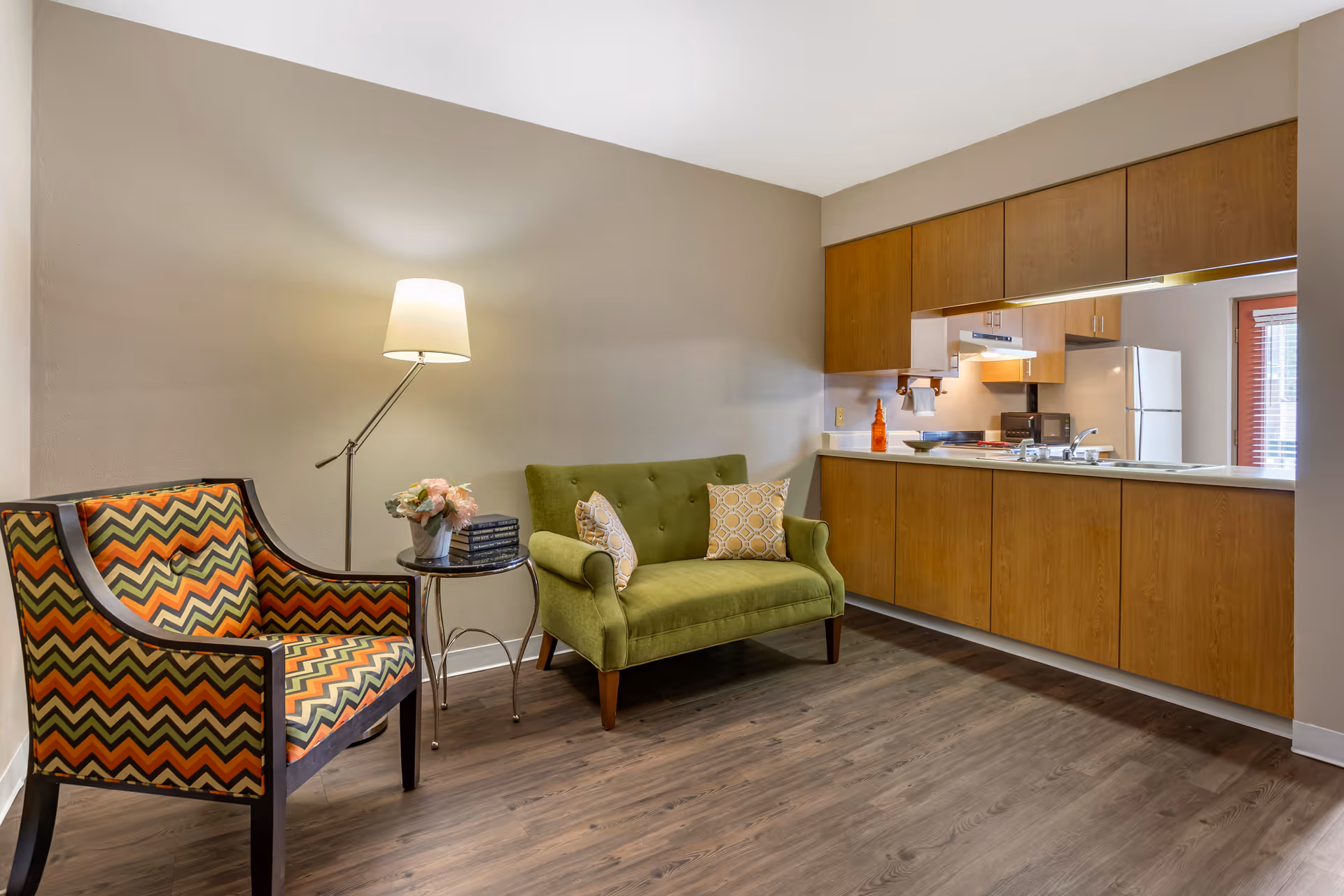Small living area with a green loveseat and patterned armchair beside a floor lamp, opening to a kitchen with wooden cabinets.