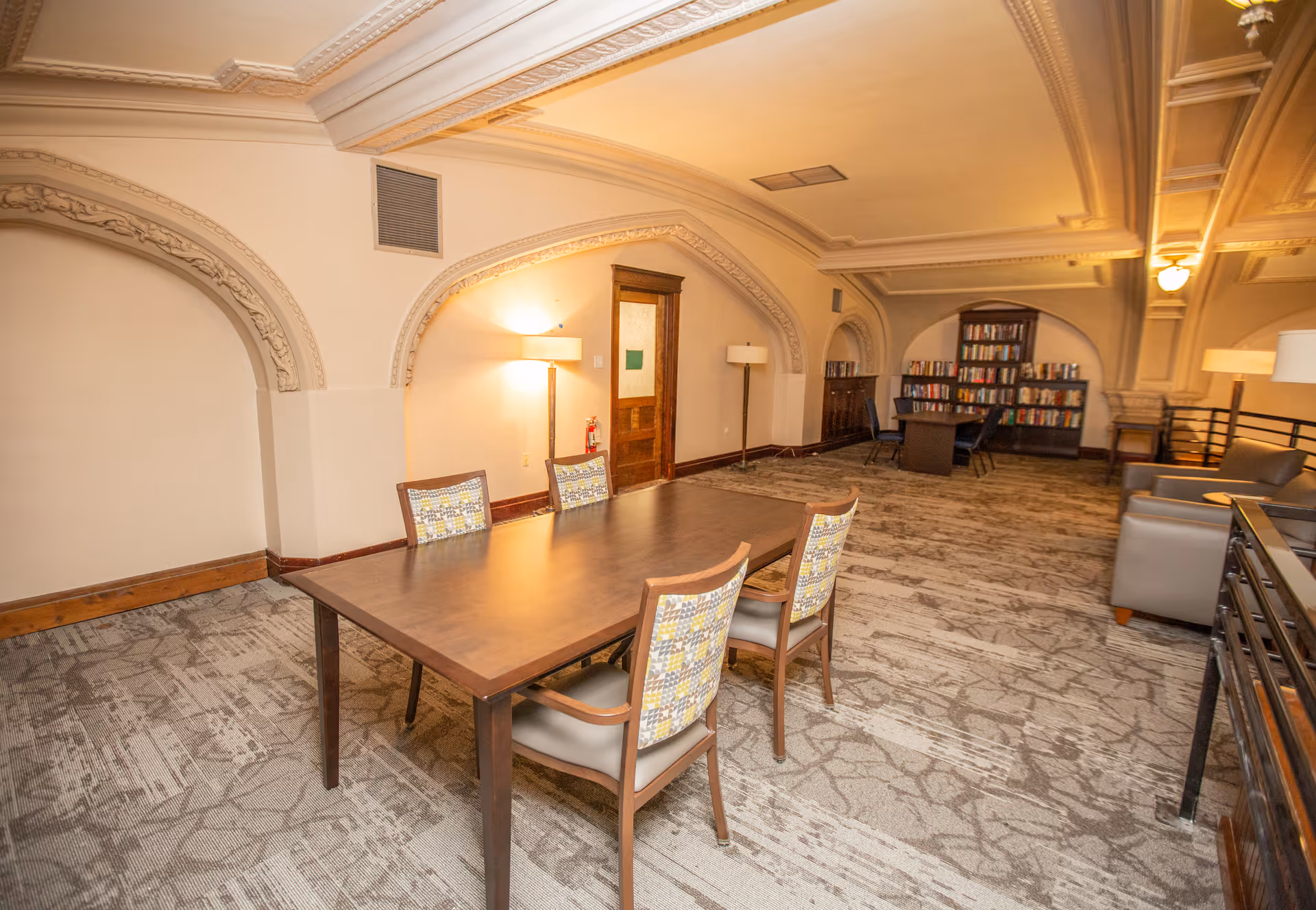 A spacious senior living common area with a large wooden table surrounded by four chairs in the foreground. The room features ornate arched wall details, carpeted floors, and warm lighting from floor lamps. In the background, there is a bookshelf filled with books and additional seating including a sofa and chairs.