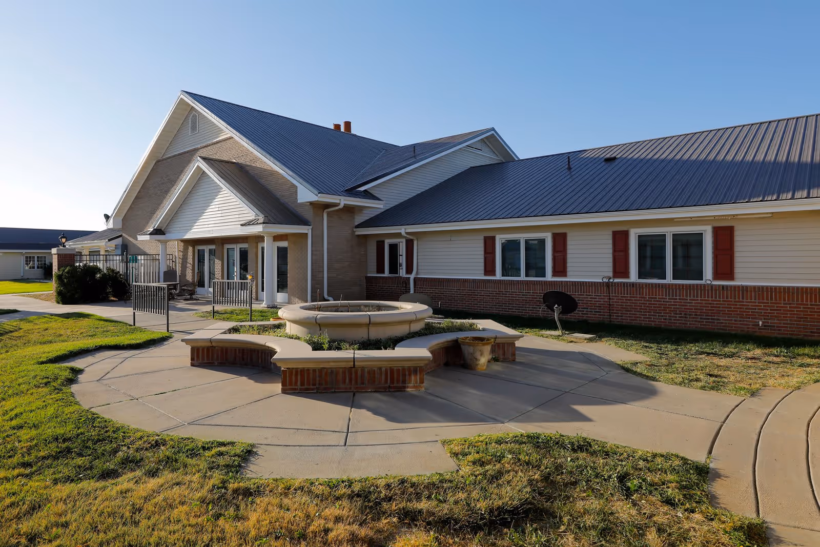 Exterior view of a senior living facility building with a metal roof, brick and siding walls, and red window shutters. In front of the building is a circular concrete seating area surrounding a central planter or fountain base, with a paved walkway and green grass around it.