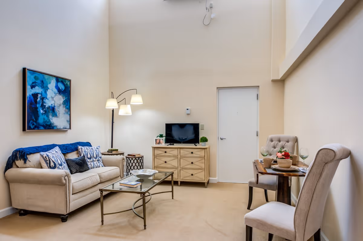 A cozy living room area with a beige sofa adorned with blue and patterned pillows, a glass coffee table, and a floor lamp with three lights. A wooden dresser with a small TV and decorative items sits against the wall, next to a closed white door. To the right, there is a small round dining table set with two upholstered chairs, tableware, and a flower centerpiece. The walls are light-colored and there is a blue abstract painting hanging above the sofa.