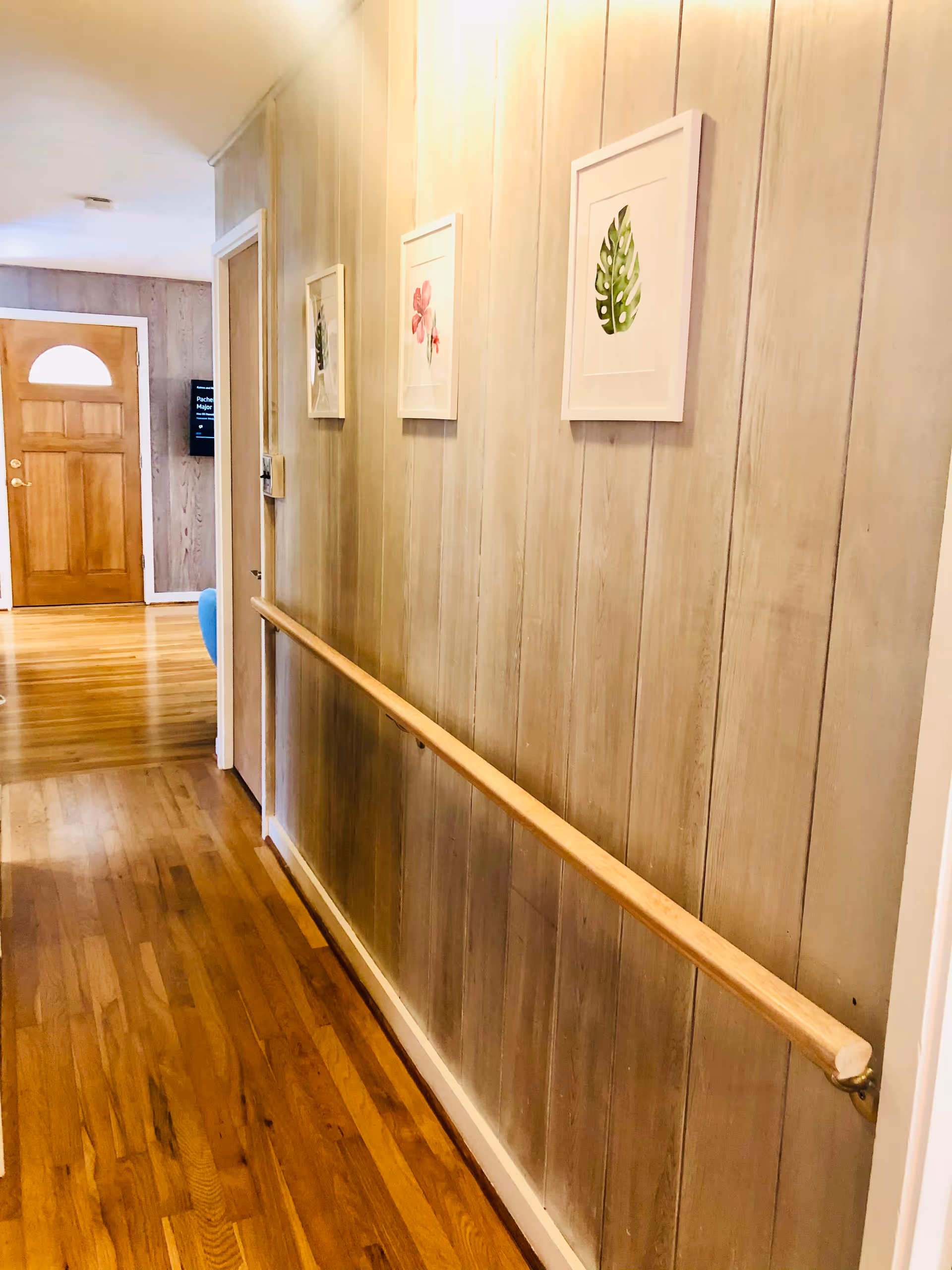 Narrow wood-paneled hallway with a wooden handrail, framed botanical prints, and a view toward a front door.