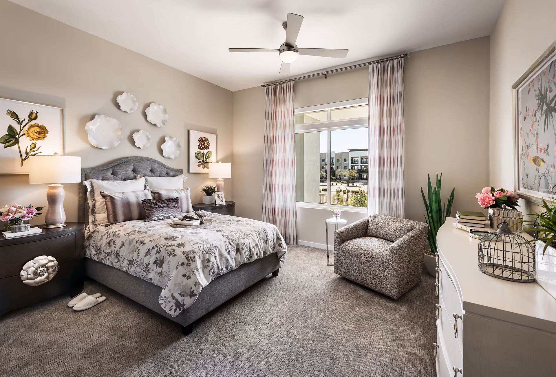 A well-lit bedroom with a gray upholstered bed adorned with floral bedding and multiple pillows. Two bedside tables with lamps flank the bed. Decorative white plates and framed floral artwork hang on the wall above the bed. A window with patterned curtains lets in natural light. A comfortable armchair and a small round side table are positioned near the window. A white dresser with books, flowers, and decorative items is on the right side of the room. The carpet is light gray, and a ceiling fan with a light is mounted on the ceiling.