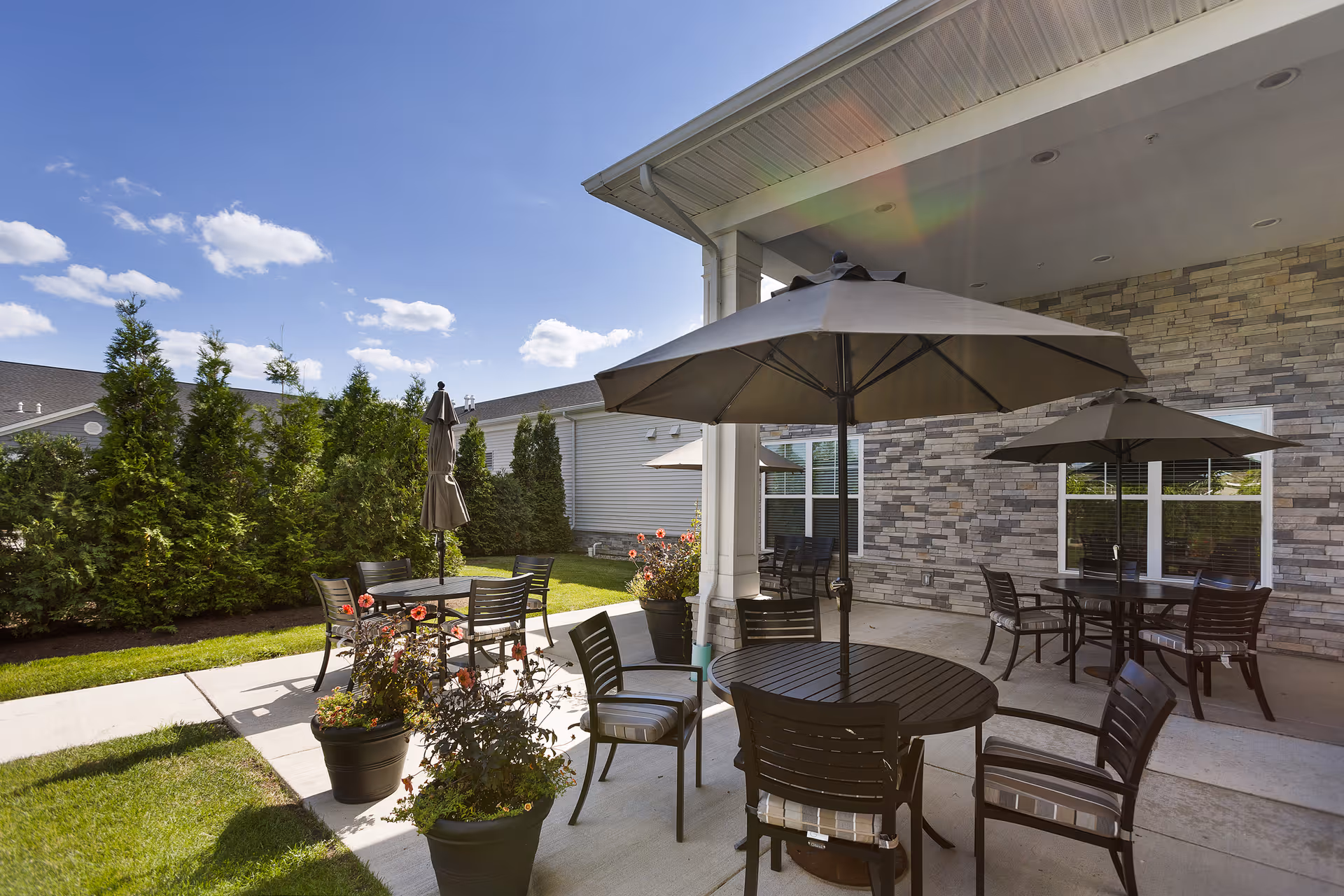 Outdoor patio area at StoryPoint Waterville with round tables, chairs, and large umbrellas. The patio is adjacent to a building with stone siding and windows. There are potted plants with flowers and a row of tall evergreen trees along the edge of the grassy lawn under a clear blue sky with some clouds.