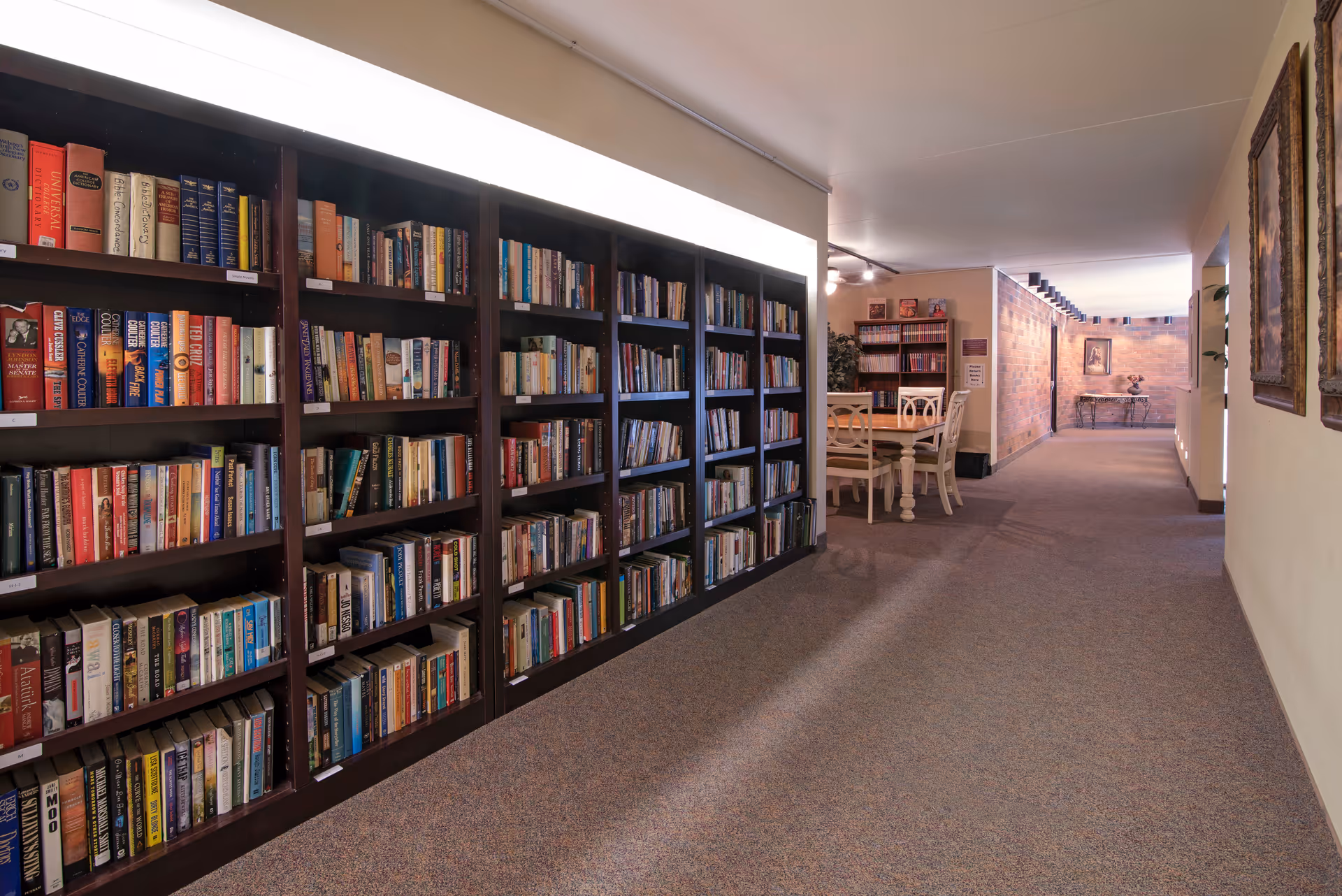 A well-lit hallway with a large bookshelf filled with books on the left side. At the end of the hallway, there is a table with chairs and more bookshelves against the wall. The walls are decorated with framed artwork and the floor is carpeted.