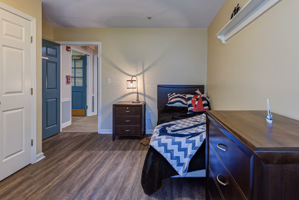 Small furnished bedroom with a single bed, nightstand and dresser, and an open doorway to a hallway.