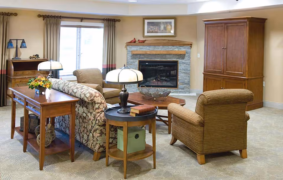 A cozy living room in a senior living facility featuring a floral patterned sofa, two upholstered armchairs, a wooden coffee table, and side tables with lamps. There is a stone fireplace with a wooden mantel and a framed picture above it. The room has beige walls, carpeted floor, and a large window with curtains.