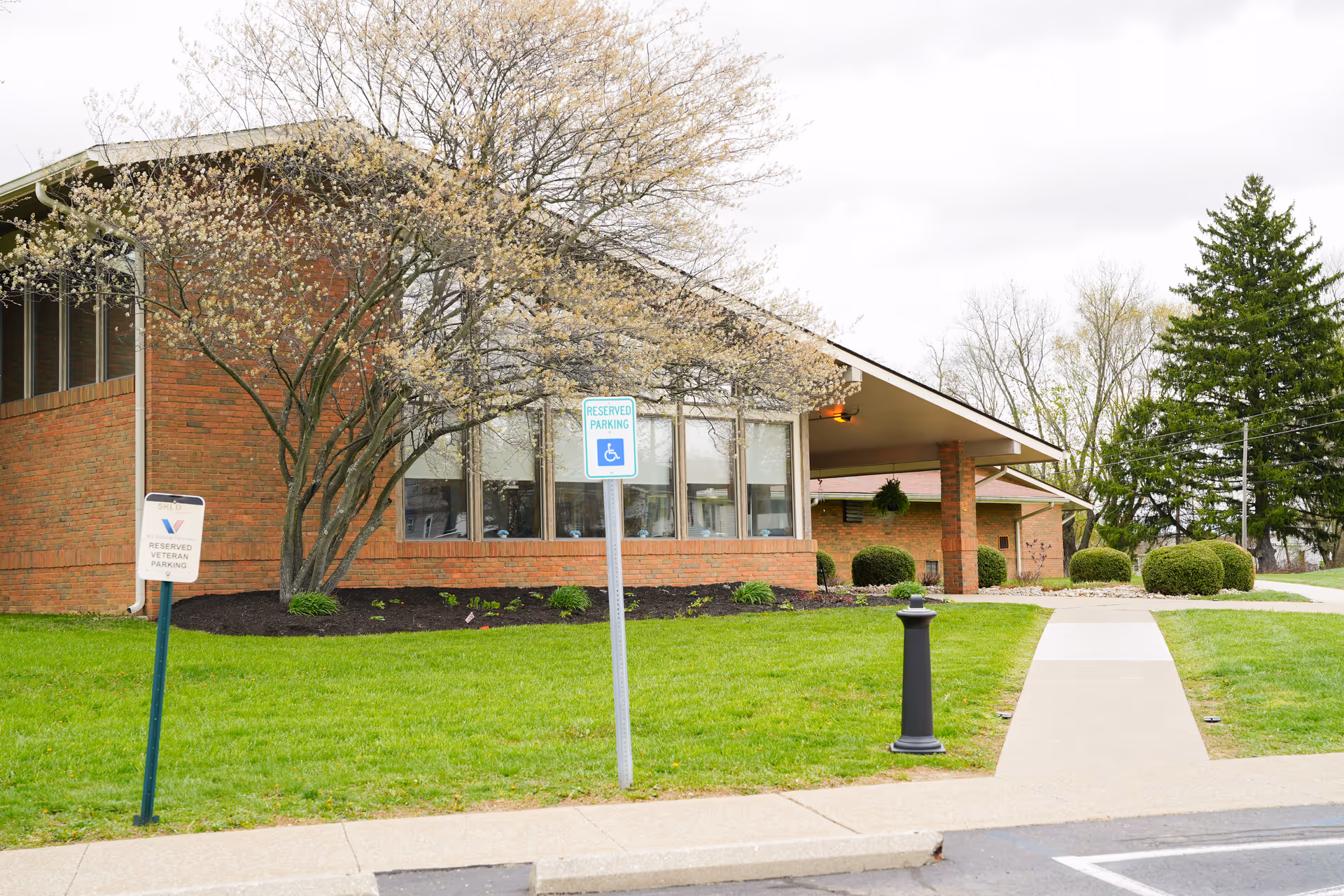 Exterior view of a brick building with large windows and a slanted roof, surrounded by green grass, bushes, and a tree with light-colored blossoms. There are two parking signs in front, one indicating reserved parking for veterans and the other for handicapped parking. A sidewalk leads to the entrance of the building.