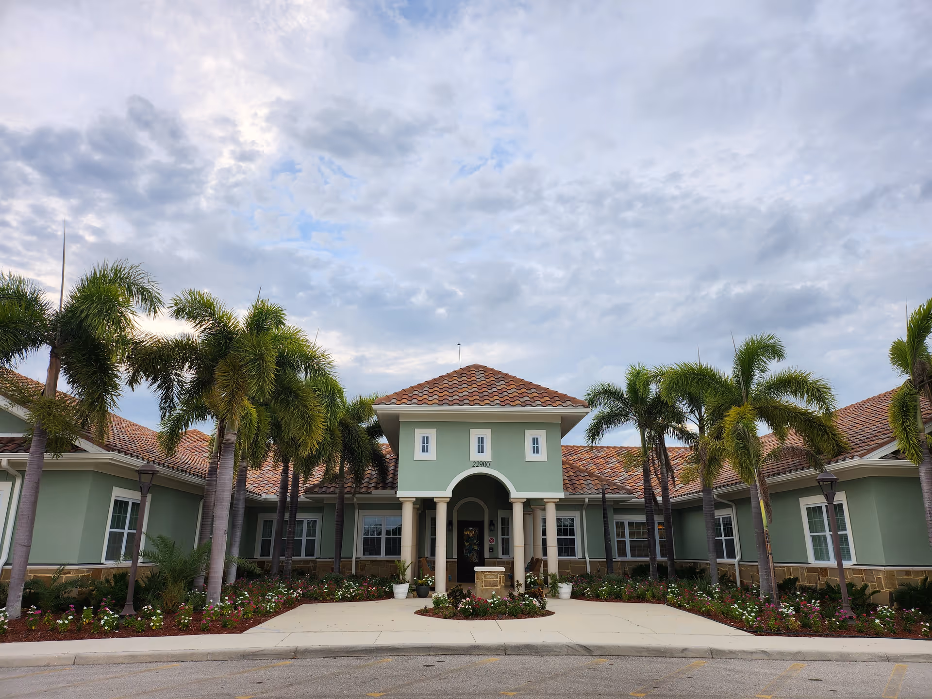 Front exterior view of Gulf Coast Memory Care facility with a tiled roof, green walls, palm trees, and landscaped flower beds under a cloudy sky.