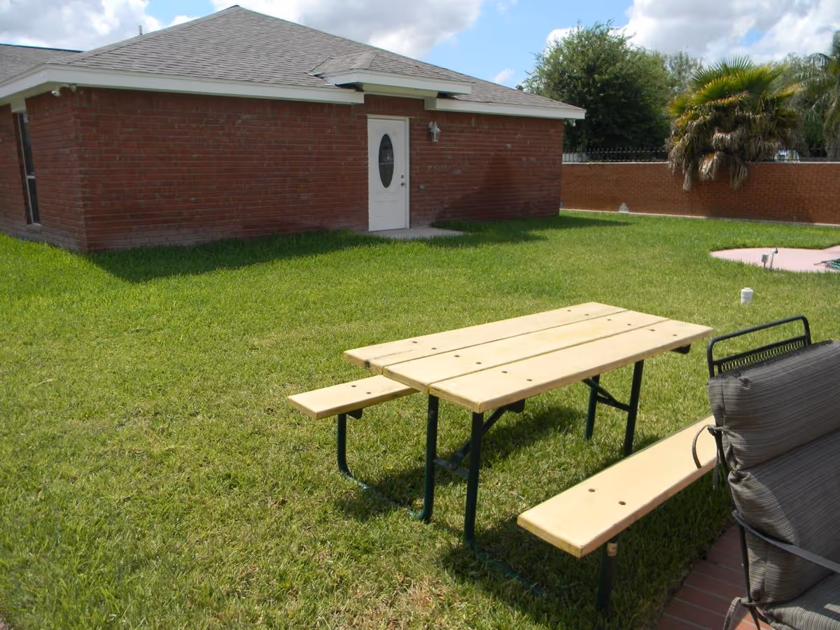 Outdoor area with a wooden picnic table and bench on a grassy lawn next to a brick building with a white door. There are trees and a brick wall in the background under a partly cloudy sky.