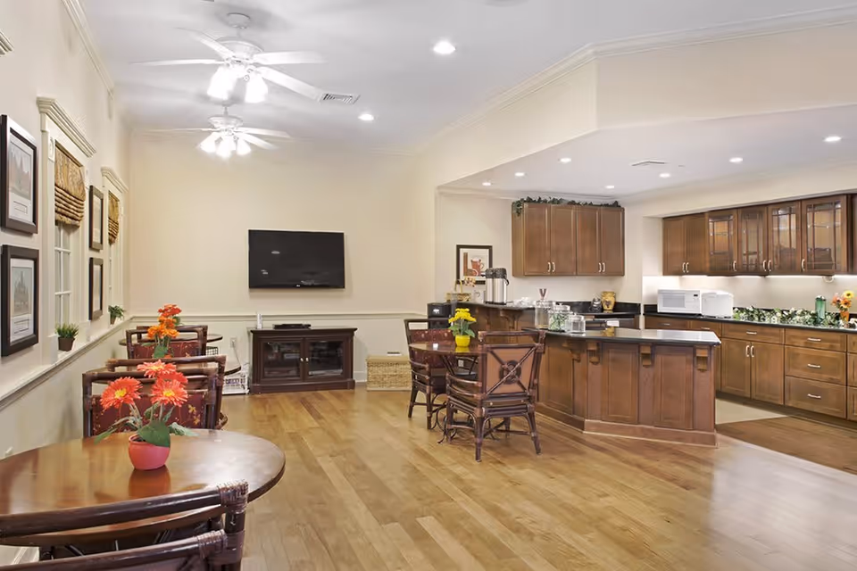 A spacious and well-lit common area in a senior living facility featuring a kitchen with wooden cabinets, a microwave, and a coffee station. There are several small round tables with chairs and flower centerpieces, a wall-mounted flat-screen TV, framed pictures on the wall, and ceiling fans with lights.