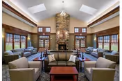 Spacious communal living room with multiple armchairs and sofas arranged around a central coffee table facing a stone fireplace under a high ceiling with skylights.
