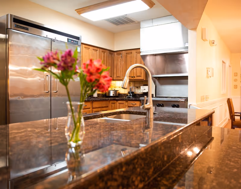 A kitchen area with a large stainless steel refrigerator, wooden cabinets, a stove with a stainless steel hood, and a granite countertop with a built-in sink and a modern faucet. A vase with pink and red flowers is placed on the countertop.