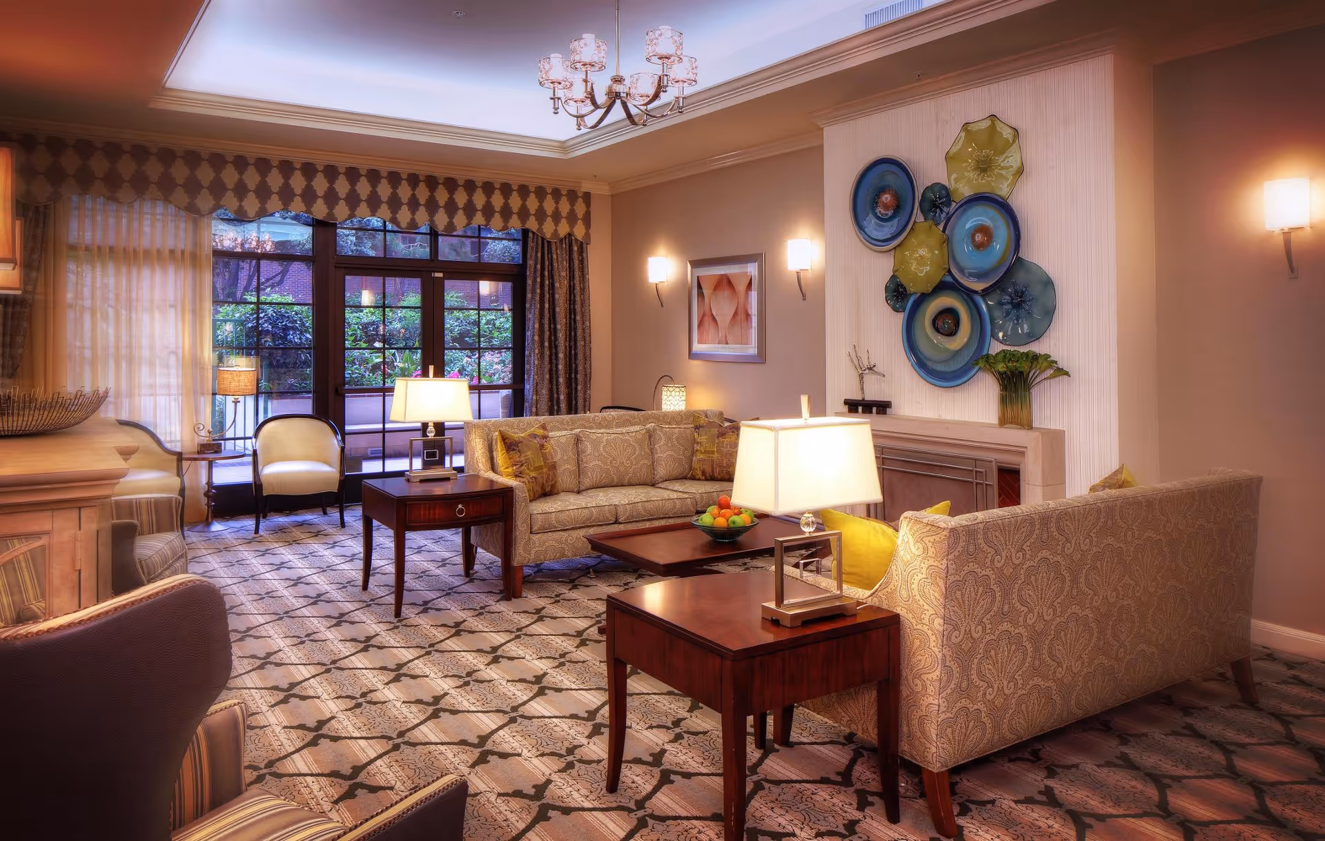 A warmly lit living room with patterned carpet, two beige sofas with decorative pillows, wooden side tables with lamps, a coffee table with a bowl of fruit, wall art featuring colorful circular designs, a framed picture, and large windows with curtains overlooking greenery outside.