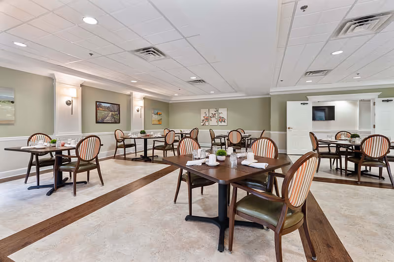 A spacious dining room with several dark wood tables and striped cushioned chairs arranged neatly. The room has light-colored walls with decorative wall panels and paintings, a tiled floor with wood accents, and recessed ceiling lights providing bright illumination.