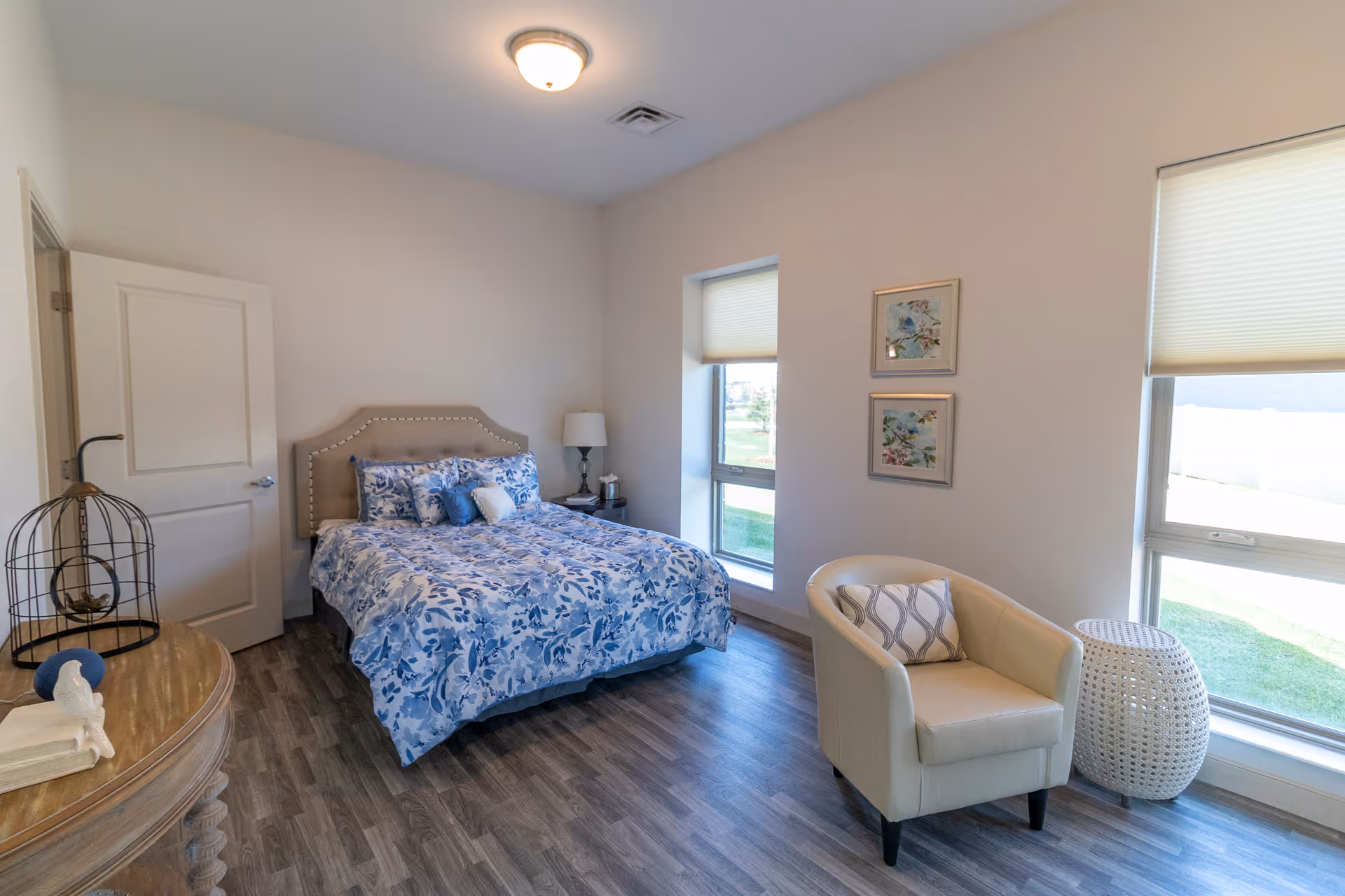 A bedroom with a bed featuring a blue and white floral comforter and pillows, a beige upholstered headboard, a wooden nightstand with a lamp and tissue box, two windows with shades partially drawn, a beige armchair with a patterned pillow, a white woven side table, and a wooden dresser with decorative items on top. The room has wood flooring and light-colored walls.