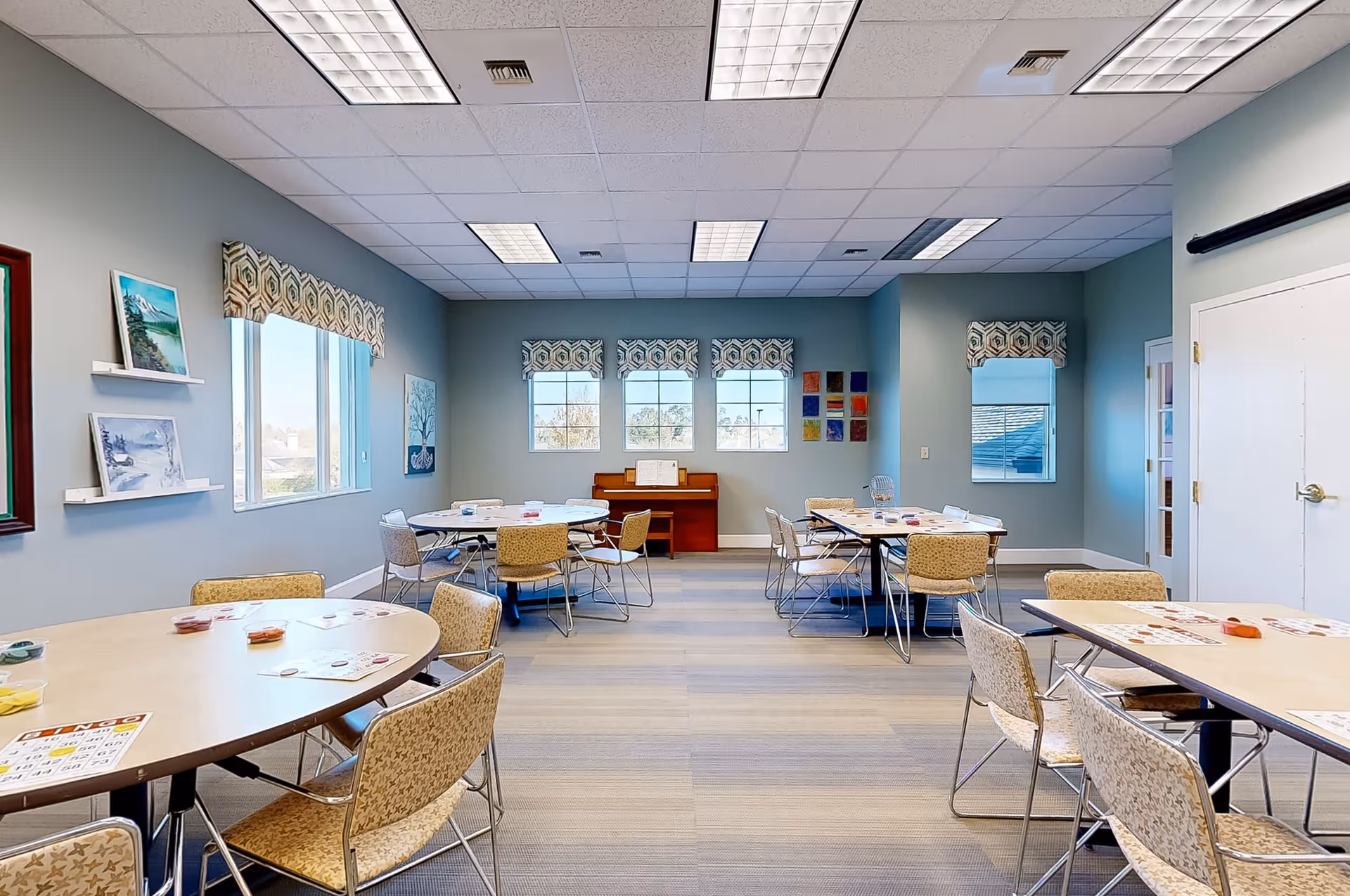 A bright, spacious activity room with multiple round and rectangular tables surrounded by chairs. The tables have bingo cards and small containers on them. The walls are painted light blue and decorated with framed pictures and colorful artwork. There are several windows with patterned valances letting in natural light, and a piano is positioned against the far wall.
