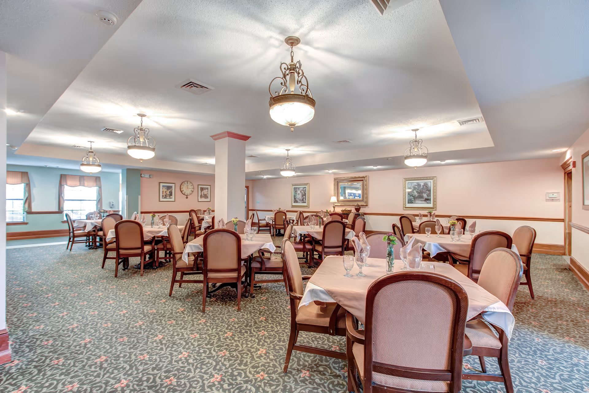 A spacious dining room with multiple tables covered with light pink tablecloths, each set with glassware, napkins, and small flower vases. The room has patterned carpet flooring, soft pink walls with framed artwork, and several hanging light fixtures providing warm lighting. Windows with valances are visible on the left side.
