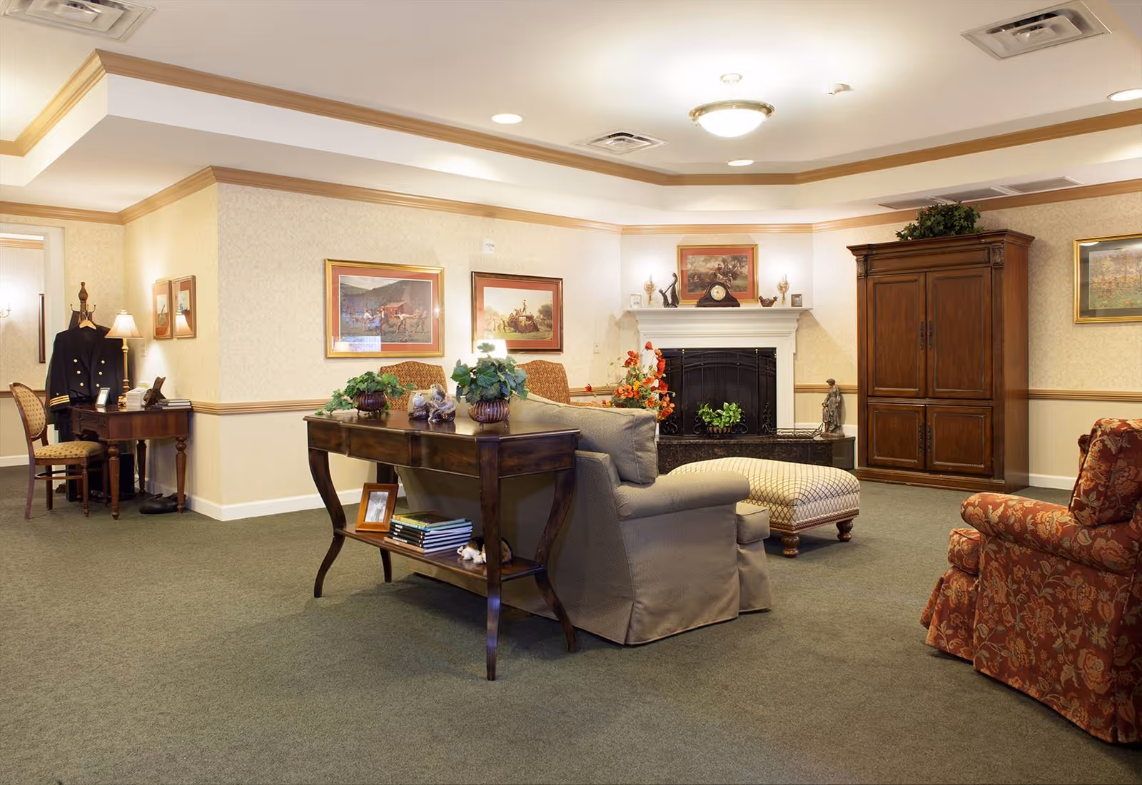 A cozy living room in a senior living facility featuring a fireplace with decorative items on the mantel, a wooden armoire, a patterned armchair, a cushioned ottoman, and a sofa with a wooden console table behind it holding plants and books. The walls are adorned with framed paintings and the room is softly lit with ceiling lights and wall sconces.