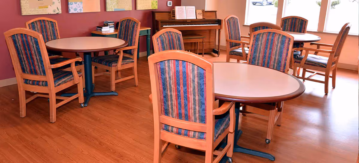 Common room with round dining tables and upholstered wooden chairs on a hardwood floor and a piano against the wall.