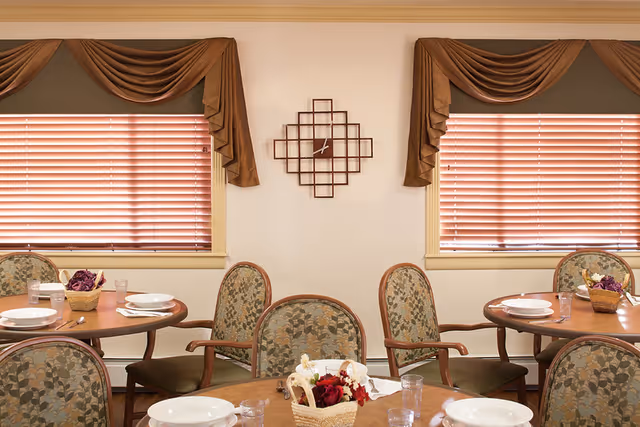 Dining room with round wooden tables set for meals, upholstered chairs, floral centerpieces, and windows with blinds and valances.