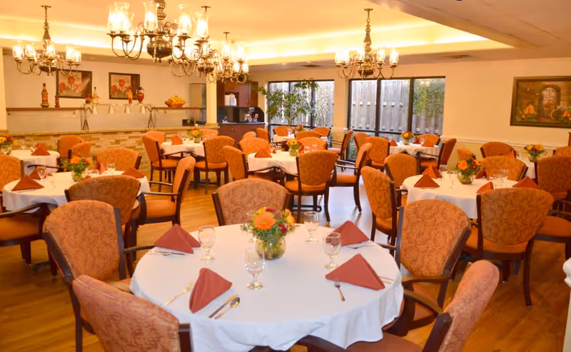 Well-lit dining room with round tables set with white tablecloths, folded napkins, glassware, floral centerpieces and upholstered chairs.