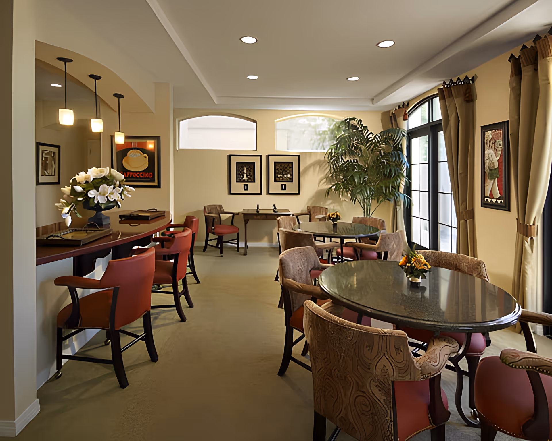 Well-lit dining/lounge room with round tables, upholstered chairs, a counter with bar stools, and large windows with curtains.