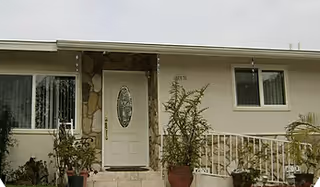 Single-story home's front exterior showing an oval-glass entry door, two windows, potted plants, stone accents, and a white railing.