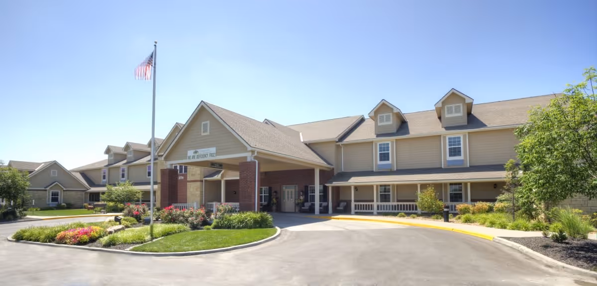 Exterior view of a senior living facility building with a covered entrance, an American flag on a flagpole, and landscaped greenery including bushes and flowers under a clear blue sky.
