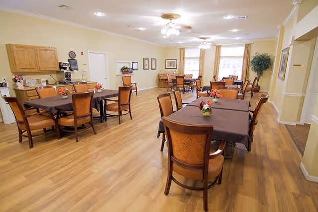 A bright dining room with multiple tables covered in brown tablecloths, wooden chairs, floral centerpieces, and large windows.