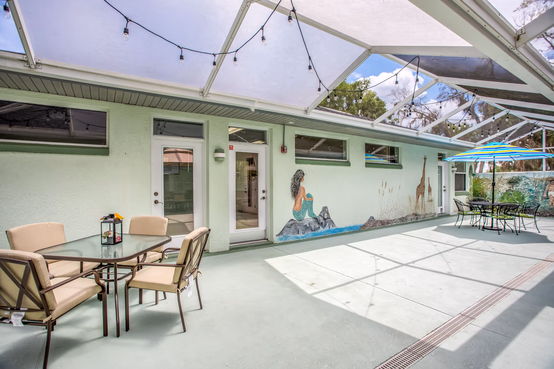 Covered outdoor courtyard with patio tables and chairs, string lights, and a mural of a mermaid and giraffes on the building wall.