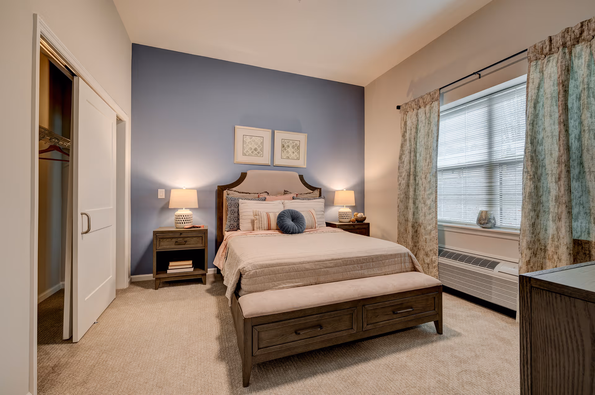 A cozy bedroom with a bed centered against a blue accent wall. The bed has a beige upholstered headboard, multiple pillows, and a beige bedspread. There are two matching wooden nightstands on either side of the bed, each with a lamp. To the left is a closet with sliding white doors, and to the right is a window with patterned curtains and a heating/cooling unit below it. The room has beige carpeting and neutral-colored walls.