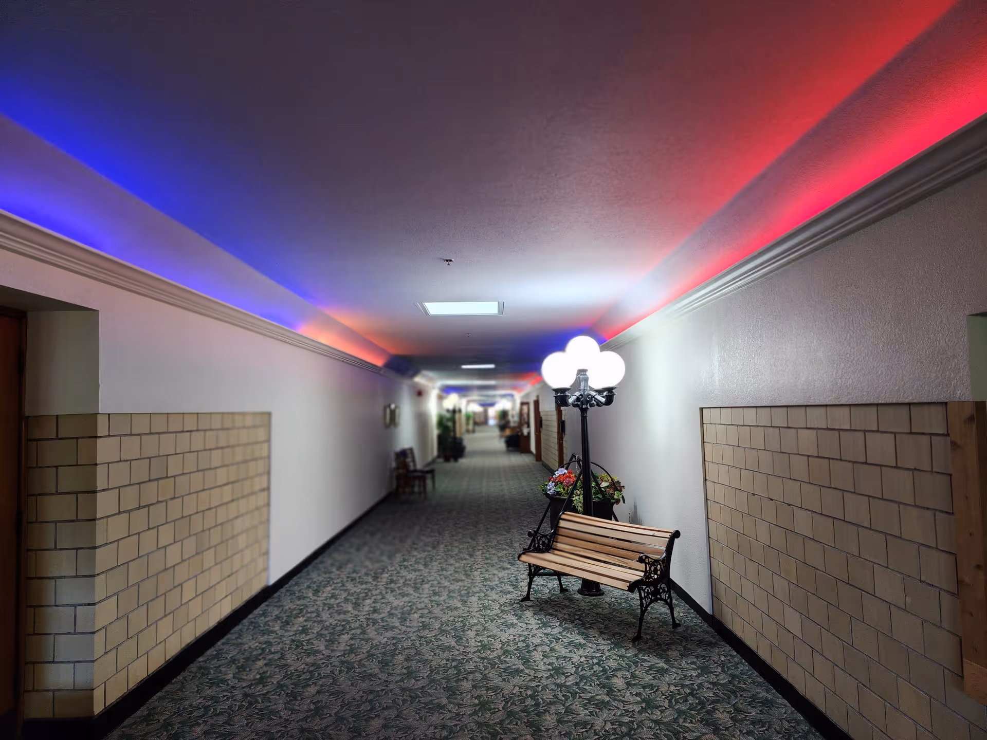 A long indoor hallway with patterned carpet and white walls partially covered with beige tiles. The ceiling has colorful lighting with blue and red hues. There are wooden benches and a vintage-style lamp post with multiple glowing white globes along the hallway. Some plants and additional benches are visible further down the corridor.