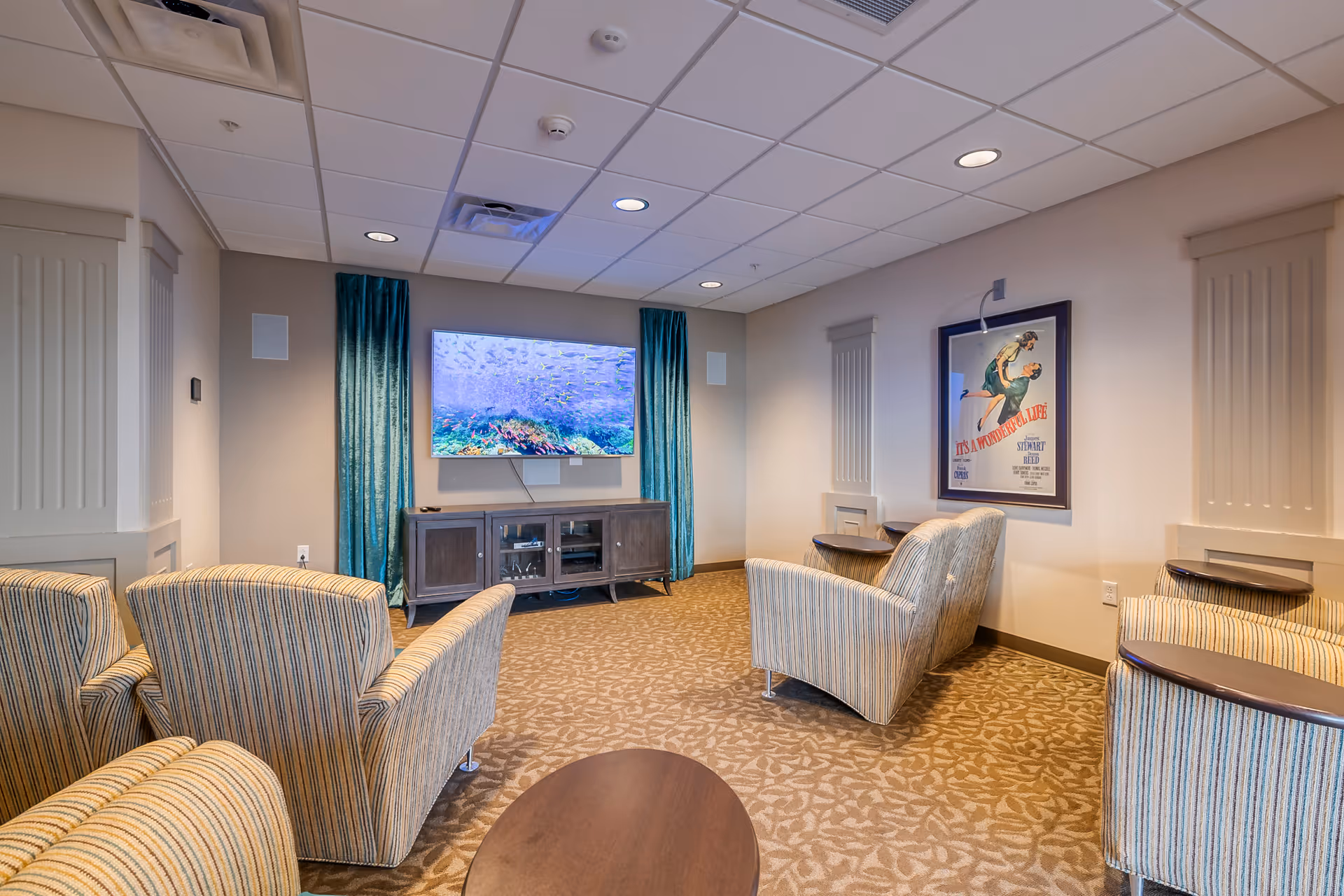 Cozy lounge with striped armchairs arranged facing a wall-mounted TV and media cabinet in a senior living facility.