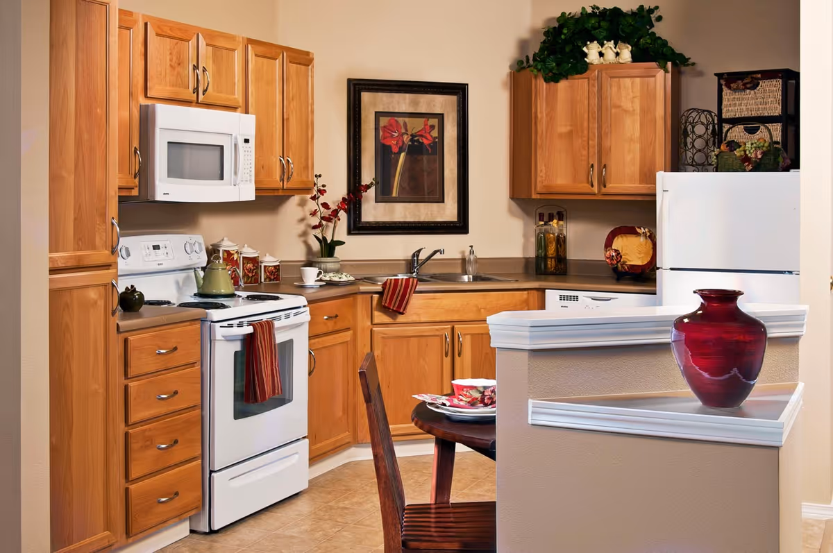 Bright kitchen with wooden cabinets, white appliances, a small dining table, and a red decorative vase.