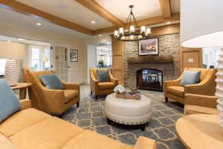 A cozy living room with beige armchairs and a matching sofa arranged around a round ottoman on a patterned rug. The room features a stone fireplace with a framed picture above it, wooden ceiling beams, and soft lighting from a chandelier and table lamps.