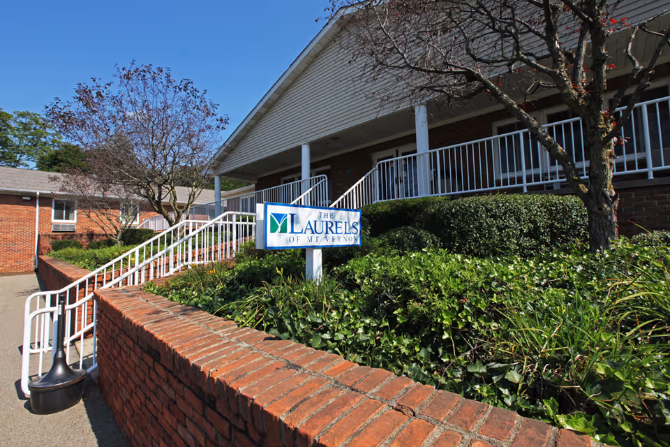 Exterior view of The Laurels of Mt. Vernon facility showing a brick building with white railings, a ramp, trees, and green shrubbery under a clear blue sky.