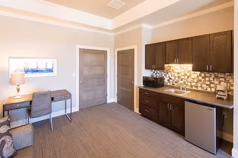 A well-lit room featuring a small kitchenette with dark wood cabinets, a sink, a mini refrigerator, a microwave, and a coffee maker. There is a desk with a chair and a lamp on the left side of the room, beige walls, carpeted floor, and two closed wooden doors.