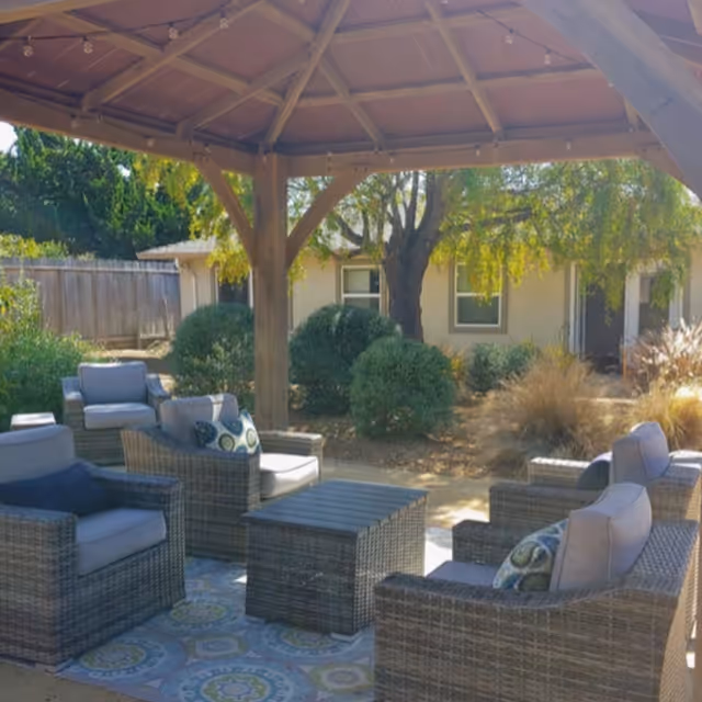 Outdoor covered patio with wicker lounge chairs and coffee tables arranged under a wooden gazebo in a landscaped courtyard.