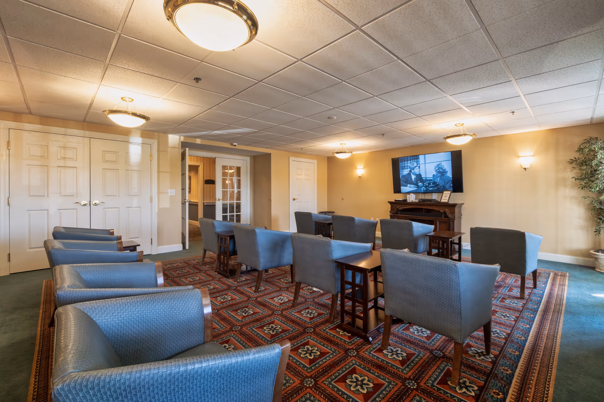 A cozy common area with multiple blue upholstered armchairs arranged in rows facing a wall-mounted flat screen TV above a wooden fireplace. The room has a patterned red and blue carpet, beige walls, ceiling lights, and a potted plant in the corner near a window letting in natural light.