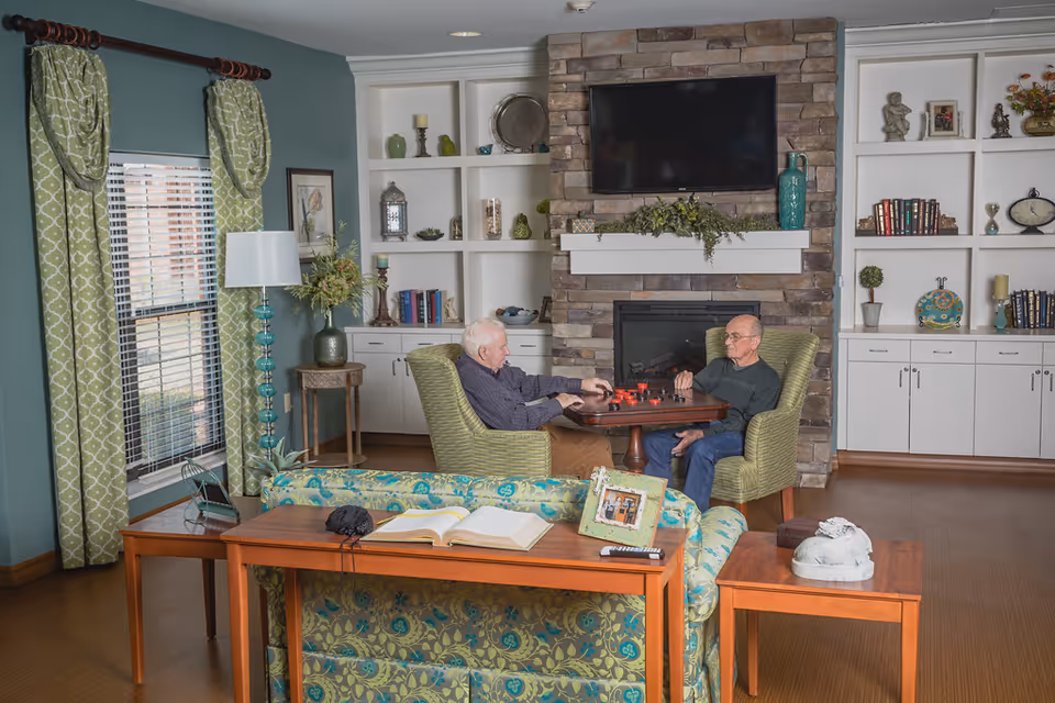 Two elderly men sitting in green armchairs playing a board game in a cozy living room with a stone fireplace, built-in white shelves filled with books and decorative items, a flat-screen TV mounted above the fireplace, green patterned curtains on the window, and a wooden table with a floral-patterned couch in the foreground.