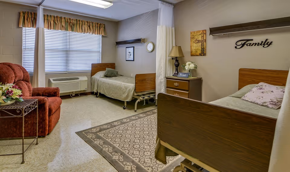 A cozy room in a senior living facility with two single beds separated by a curtain. The room features a red armchair, a small table with flowers, a dresser with a lamp and decorative items, and wall decor including a clock and the word 'Family'. A window with blinds and a valance lets in natural light.