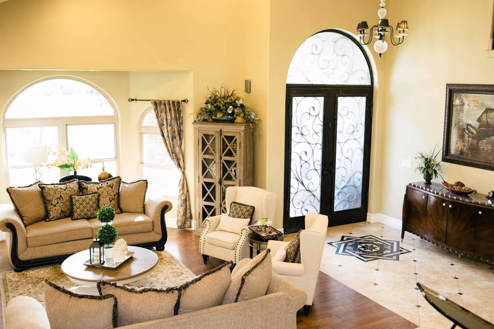 Bright elegant living room with sofas and armchairs arranged around a coffee table near an ornate double-door entry.