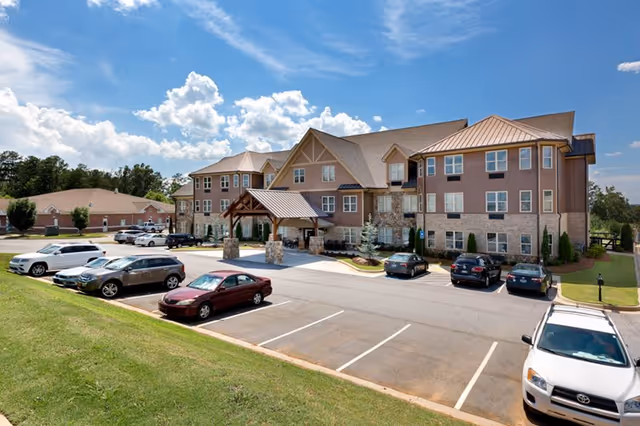 Front exterior of a three-story senior living building with a covered entrance, parking lot, and several parked cars under a blue sky.