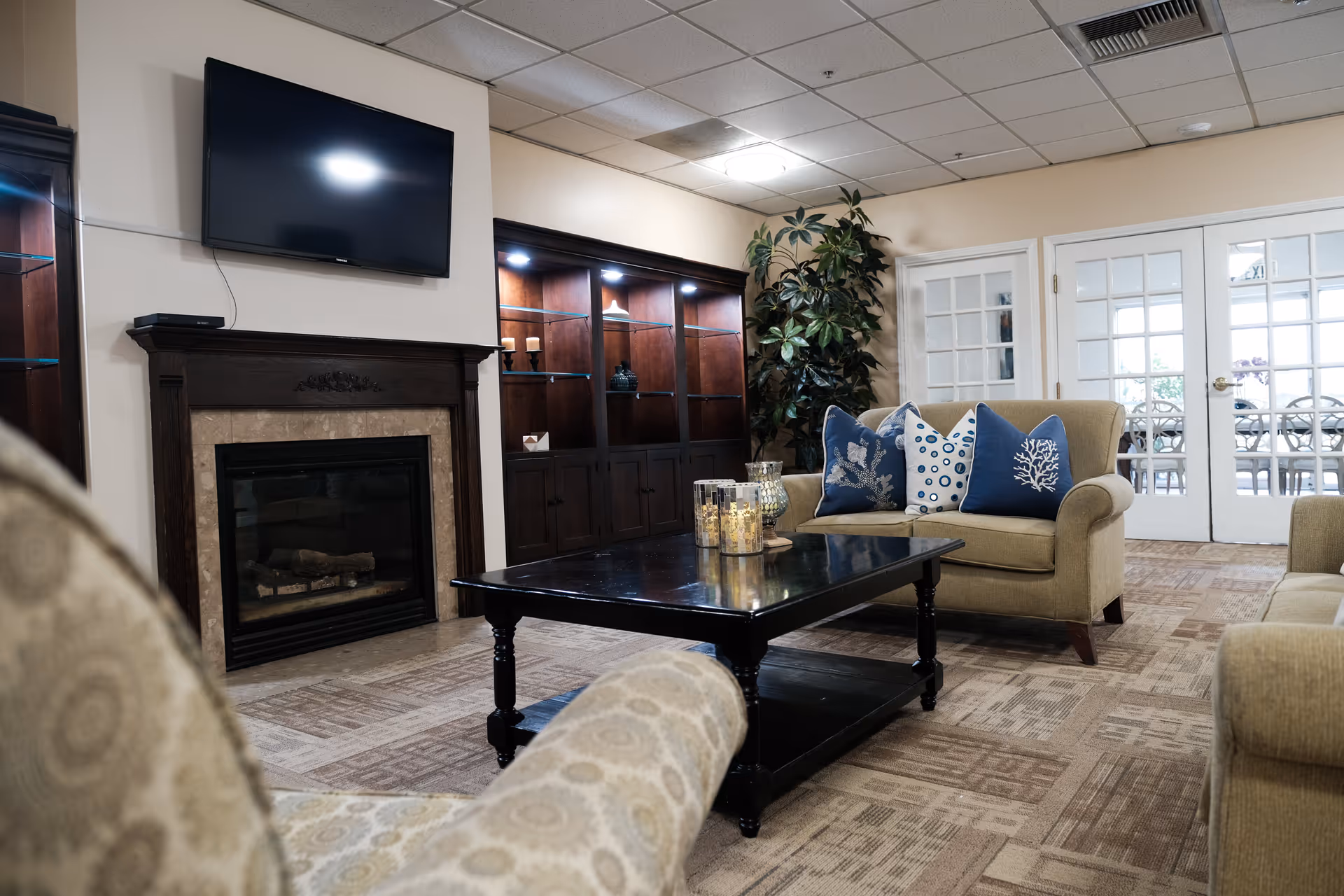 A cozy living room area with beige upholstered armchairs and a loveseat adorned with blue and white decorative pillows. A dark wooden coffee table with candles sits in the center. There is a fireplace with a mounted flat-screen TV above it, and a dark wooden cabinet with glass shelves and spotlights in the background. French doors with glass panes lead to another room.