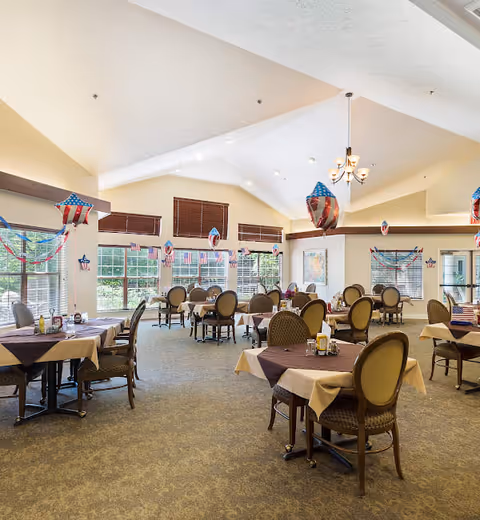 A spacious dining room decorated with American flag-themed balloons and garlands. Multiple tables are set with tablecloths, chairs, and condiments. Large windows allow natural light to fill the room, and the ceiling features a chandelier and recessed lighting.