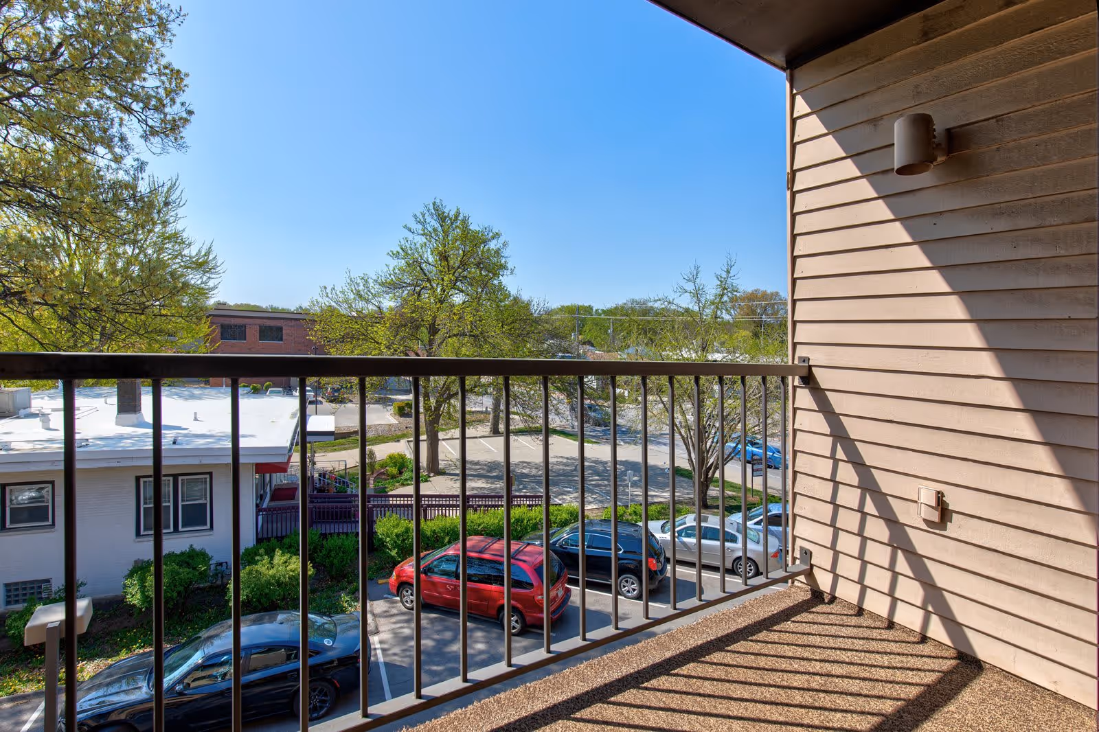 View from a balcony overlooking a parking lot with several parked cars, trees with green leaves, and nearby buildings under a clear blue sky.