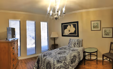 A single bed with patterned bedding is placed against a beige wall in a well-lit room. Next to the bed is a round glass-top side table and a wooden chair. On the wall above the bed are three framed artworks. A floor lamp stands in the corner near three tall windows with blinds. Across from the bed is a wooden dresser with a flat-screen TV on top. The room has wooden flooring and a chandelier hanging from the ceiling.