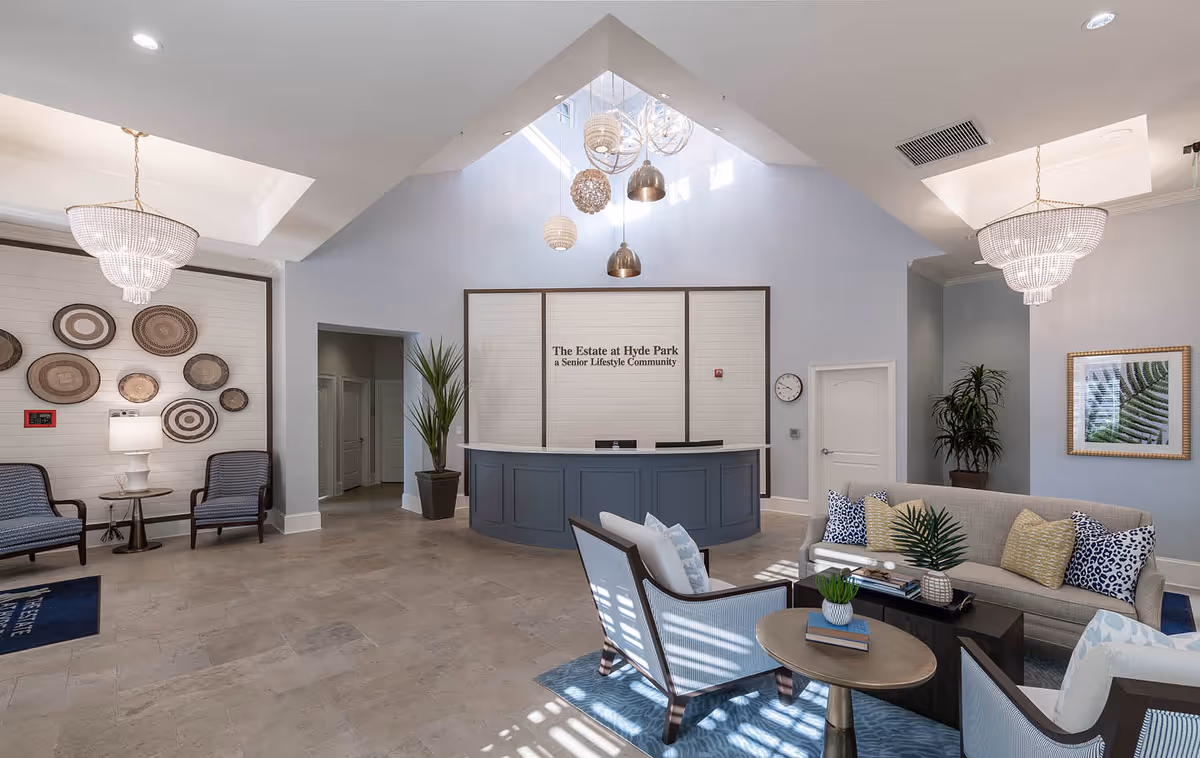 Bright and spacious senior living community reception area with a blue curved front desk under a high vaulted ceiling with hanging pendant lights. The room features comfortable seating including a beige sofa with patterned pillows, two armchairs, and a small round table with a plant. Decorative wall art and plants add warmth to the space.