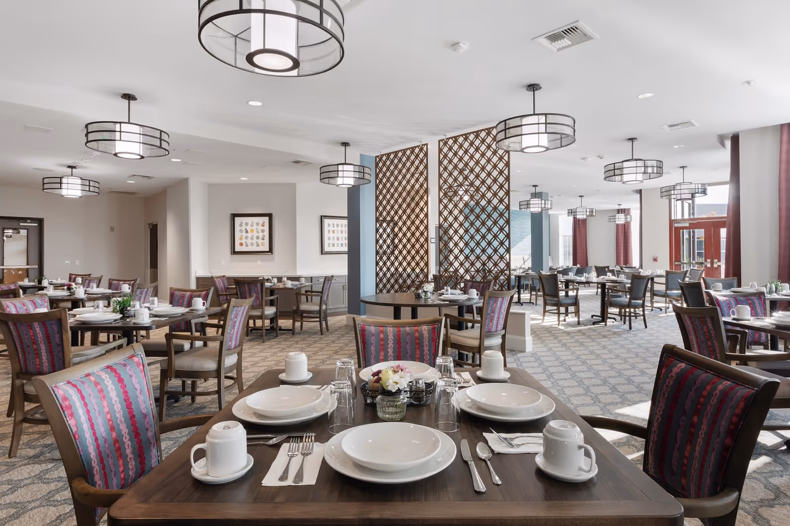 A spacious and well-lit dining room with multiple tables set for meals. Each table has white plates, cups, silverware, and small flower arrangements. The room features modern hanging light fixtures, patterned carpet, and wooden chairs with colorful upholstery. Large windows with red curtains allow natural light to fill the space.