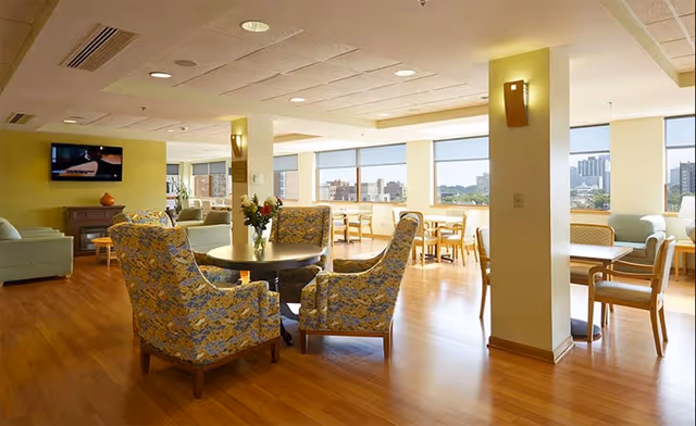 A bright and spacious senior living community lounge area with wooden flooring, several seating arrangements including floral upholstered armchairs around a round table, light green sofas, and wooden chairs with tables near large windows offering a city view. The room is well-lit with ceiling lights and wall sconces.